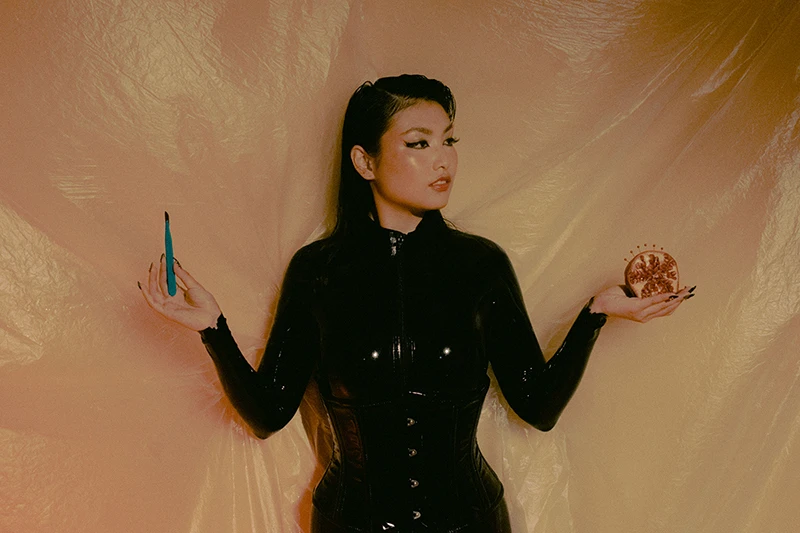 Mx Tomie standing in a black catsuit holding a scalpel in one hand and some fruit in the other