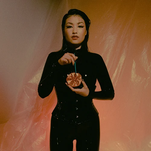 Mx Tomie wearing black latex holding a pomegranate