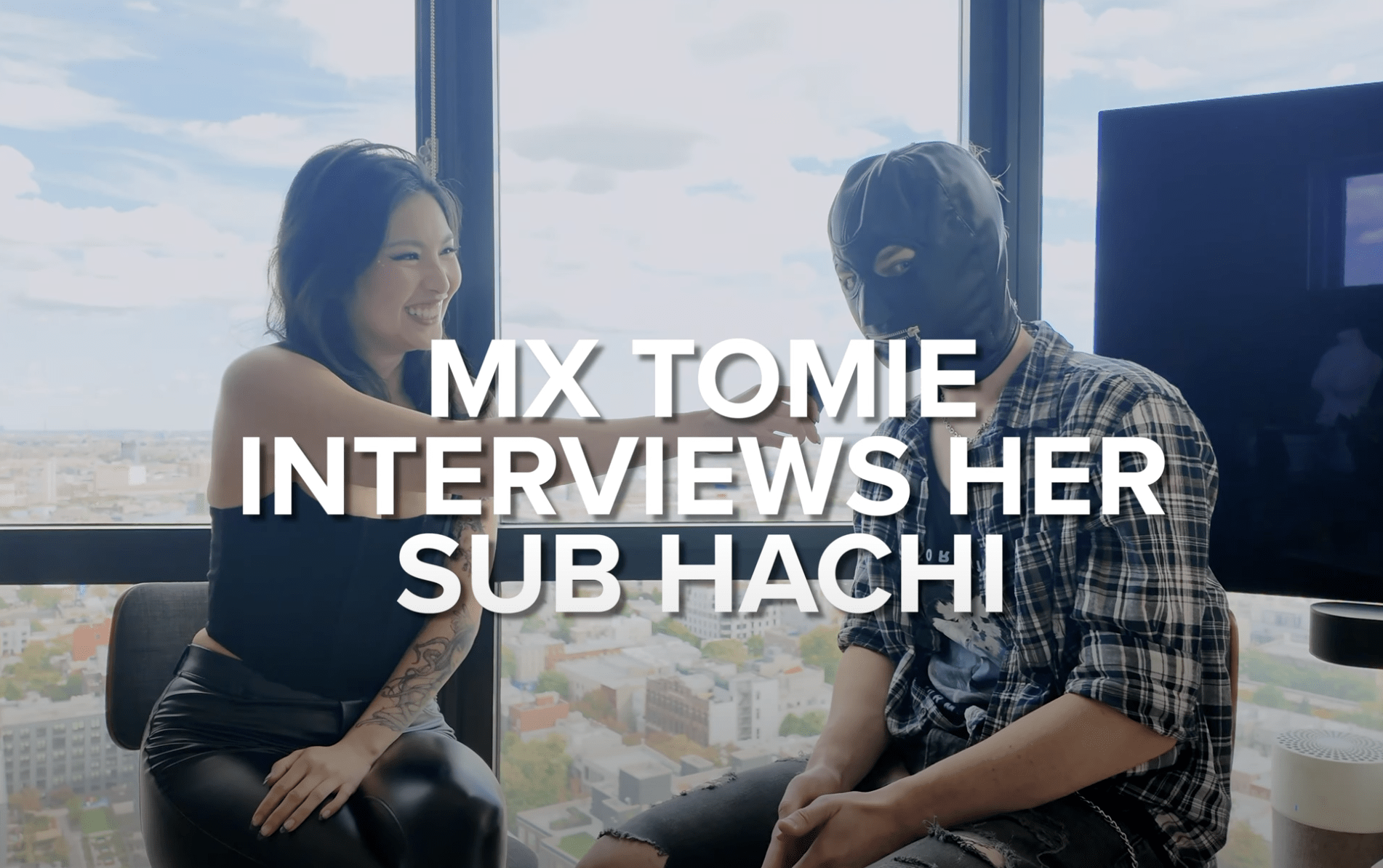 Mx Tomie interviews her sub Hachi. Hachi is wearing a hood and Mx Tomie is wearing all black holding a microphone.